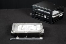 3,5"-Festplatte mit Montageschienen, dahinter ein Festplattenkäfig mit einer montierten Festplatte